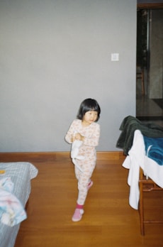 A young child wearing pajamas with a floral pattern is walking indoors, holding a stuffed toy. The room has wooden flooring and a neutral-colored wall. There is a bed with a blanket partially visible on the left and a chair with clothes draped over it on the right.