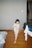 A young child wearing pajamas with a floral pattern is walking indoors, holding a stuffed toy. The room has wooden flooring and a neutral-colored wall. There is a bed with a blanket partially visible on the left and a chair with clothes draped over it on the right.