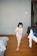 A young child wearing pajamas with a floral pattern is walking indoors, holding a stuffed toy. The room has wooden flooring and a neutral-colored wall. There is a bed with a blanket partially visible on the left and a chair with clothes draped over it on the right.