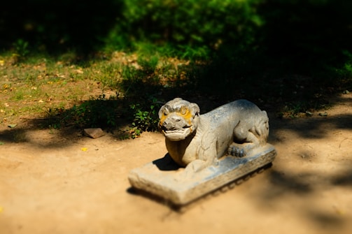 A stone sculpture of a mythical creature resembling a lion rests on a pedestal in an outdoor setting. The sculpture is weathered with time, displaying intricate carvings and a fierce expression, with distinctive yellow paint highlighting its eyes. It sits on a patch of dirt surrounded by patches of grass and shadows from nearby trees.
