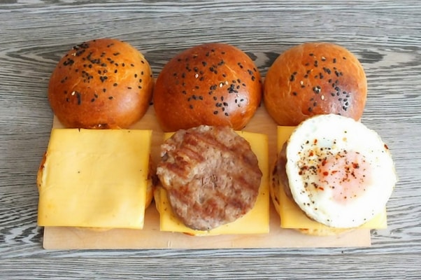 Three burger buns with sesame seeds sit on a wooden board, each paired with a slice of cheese. One bun is topped with a cooked sausage patty, and another has a fried egg seasoned with pepper and spices.