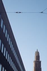 A modern cityscape features a tall building under construction with scaffolding around it. On the left, a dark, angled building displays rows of windows. Overhead, a utility cable stretches across the sky.