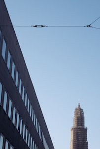 A modern cityscape features a tall building under construction with scaffolding around it. On the left, a dark, angled building displays rows of windows. Overhead, a utility cable stretches across the sky.