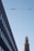 A modern cityscape features a tall building under construction with scaffolding around it. On the left, a dark, angled building displays rows of windows. Overhead, a utility cable stretches across the sky.