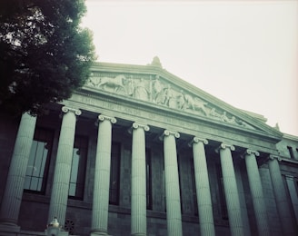 A neoclassical building facade with a row of tall columns and a triangular pediment. The pediment features intricate sculptures depicting human figures and horses. Partially visible text reads 'THE HIGHLANDS OF THE MIND LET ME GO'. A large tree is seen to the left, adding a natural element to the architecture.