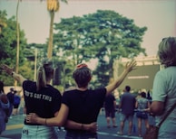 People stand in an outdoor setting, with two individuals in the foreground embracing and raising their arms. The person on the left wears a shirt with the words 'GOD'S GOT THIS'. A group of people is gathered, possibly attending an event. Trees and a large screen are visible in the background.