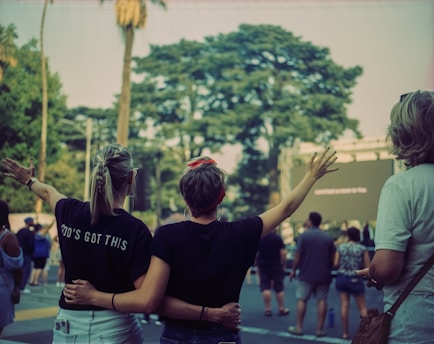 People stand in an outdoor setting, with two individuals in the foreground embracing and raising their arms. The person on the left wears a shirt with the words 'GOD'S GOT THIS'. A group of people is gathered, possibly attending an event. Trees and a large screen are visible in the background.