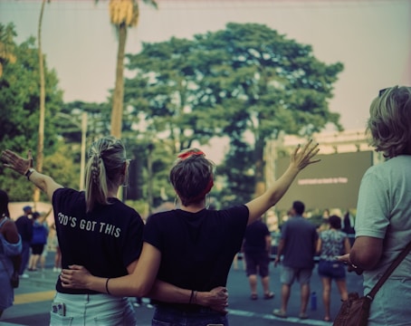 People stand in an outdoor setting, with two individuals in the foreground embracing and raising their arms. The person on the left wears a shirt with the words 'GOD'S GOT THIS'. A group of people is gathered, possibly attending an event. Trees and a large screen are visible in the background.