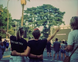 People stand in an outdoor setting, with two individuals in the foreground embracing and raising their arms. The person on the left wears a shirt with the words 'GOD'S GOT THIS'. A group of people is gathered, possibly attending an event. Trees and a large screen are visible in the background.