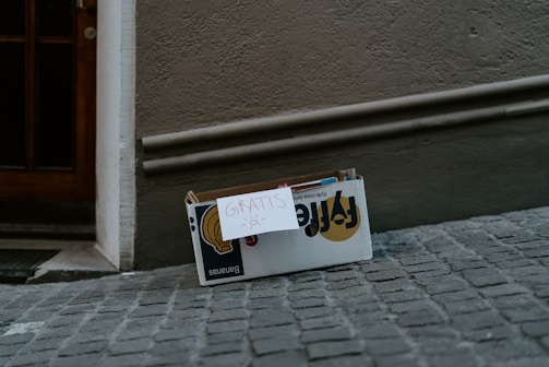 A cardboard box is placed on a cobblestone street against a textured wall. A handwritten sign labeled 'Gratis' with a small drawing of a sun is attached to the box. Inside the box are some books or magazines with partly visible covers.