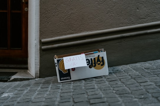 A cardboard box is placed on a cobblestone street against a textured wall. A handwritten sign labeled 'Gratis' with a small drawing of a sun is attached to the box. Inside the box are some books or magazines with partly visible covers.