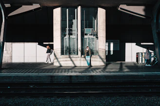 A train station platform with three people present. One person is in the center, leaning against a structure, talking on a phone. Another individual to the left is standing and looking at their phone. A third person is on the right with a stroller, appearing to be waiting. Shadows are cast on the floor from the overhead structures, creating an interplay of light and shadow.