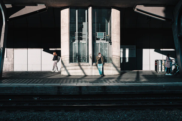 A train station platform with three people present. One person is in the center, leaning against a structure, talking on a phone. Another individual to the left is standing and looking at their phone. A third person is on the right with a stroller, appearing to be waiting. Shadows are cast on the floor from the overhead structures, creating an interplay of light and shadow.