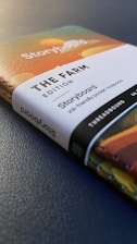 A close-up view of a notebook titled 'The Farm Edition Storyboard'. The cover features colorful abstract designs, and the notebook is thread-bound with a visible spine. The words are printed clearly, and the notebook is resting on a textured, dark surface.