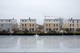 A row of modern residential buildings is reflected in a large, smooth icy surface that resembles a frozen pond or canal. The buildings have a minimalist style with flat roofs and are painted in neutral tones like beige, grey, and white. Some of the lawns also have patches of snow. A construction crane is visible in the background, indicating ongoing development in the area. Ladders are placed at the edge of the icy surface, suggesting access to the ice.