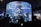 A large cylindrical aquarium filled with a variety of colorful fish and marine life is surrounded by several silhouettes of people observing. The aquarium is well-lit, showcasing the aquatic environment with rocks and plants, while the background is dimly lit, adding contrast.