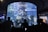 A large cylindrical aquarium filled with a variety of colorful fish and marine life is surrounded by several silhouettes of people observing. The aquarium is well-lit, showcasing the aquatic environment with rocks and plants, while the background is dimly lit, adding contrast.
