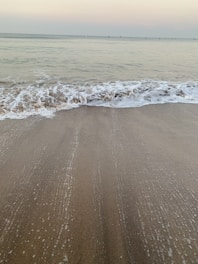 A calm shoreline at sunset with gentle waves lapping the sand under muted green and blue skies.