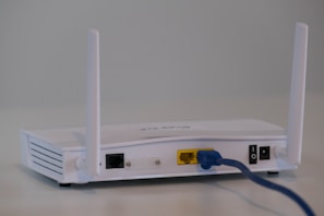 A white wireless router with two antennas is placed on a flat surface. The back of the router is visible, showing various ports including a yellow Ethernet port with a blue cable connected, power input, and a power switch.