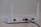 A white wireless router with two antennas is placed on a flat surface. The back of the router is visible, showing various ports including a yellow Ethernet port with a blue cable connected, power input, and a power switch.