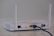 A white wireless router with two antennas is placed on a flat surface. The back of the router is visible, showing various ports including a yellow Ethernet port with a blue cable connected, power input, and a power switch.