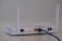 A white wireless router with two antennas is placed on a flat surface. The back of the router is visible, showing various ports including a yellow Ethernet port with a blue cable connected, power input, and a power switch.