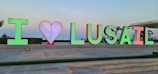 Colorful letters spell 'I ♥ LUSAIL' against an outdoor backdrop. The heart symbol is depicted in red between the initial letter 'I' and the word 'LUSAIL.' People are visible in the background, suggesting a lively public area. The sky is clear, contributing to a serene atmosphere.