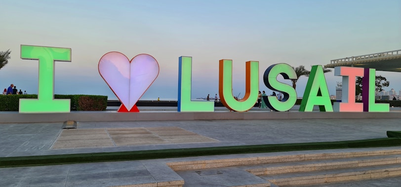 Colorful letters spell 'I ♥ LUSAIL' against an outdoor backdrop. The heart symbol is depicted in red between the initial letter 'I' and the word 'LUSAIL.' People are visible in the background, suggesting a lively public area. The sky is clear, contributing to a serene atmosphere.