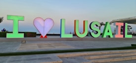 Colorful letters spell 'I &hearts; LUSAIL' against an outdoor backdrop. The heart symbol is depicted in red between the initial letter 'I' and the word 'LUSAIL.' People are visible in the background, suggesting a lively public area. The sky is clear, contributing to a serene atmosphere.