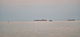 A serene seascape features a calm, expansive body of water with two large ships and a small boat in the distance, navigating between green and red buoys. The sky is overcast, casting a muted glow over the scene.