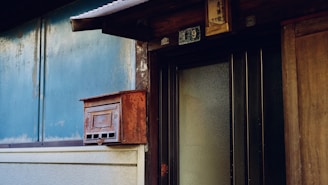 A rustic and weathered exterior wall features a rusty red mailbox mounted on a blue wall. The door nearby is made of wood with frosted glass panels, and there is a sign above it displaying the number '19'. The overall setting suggests an old, possibly abandoned or rural house.