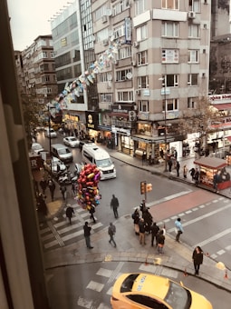A busy urban street intersection with pedestrians crossing, a cluster of colorful balloons being sold, several vehicles including a yellow taxi and a white van, and tall buildings adorned with eclectic signage. The scene features shops with vibrant signs and a festive display of flags hanging across the buildings.