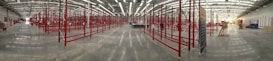 A large, empty industrial space with polished concrete floors. The area is lined with numerous red metal storage racks which span the length of the building. The ceiling is high, equipped with bright fluorescent lighting. There are a few scattered objects, including a blue lift on the right.