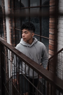 A young person is wearing a gray hoodie while walking down a staircase by a large window framed with bricks. The setting has an industrial and urban feel, with metal railings and exposed brick walls.