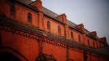 A brick facade of a historic building with multiple arched windows and decorative brickwork. The upper portion features a series of small, arched windows set within the reddish-orange brick structure. The roof is made of slate tiles, and the brickwork includes intricate patterns and designs.