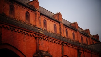 A brick facade of a historic building with multiple arched windows and decorative brickwork. The upper portion features a series of small, arched windows set within the reddish-orange brick structure. The roof is made of slate tiles, and the brickwork includes intricate patterns and designs.