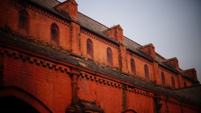 A brick facade of a historic building with multiple arched windows and decorative brickwork. The upper portion features a series of small, arched windows set within the reddish-orange brick structure. The roof is made of slate tiles, and the brickwork includes intricate patterns and designs.