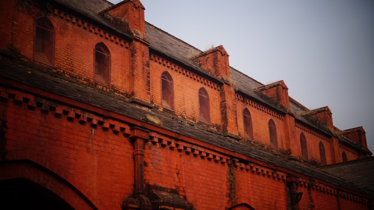 A brick facade of a historic building with multiple arched windows and decorative brickwork. The upper portion features a series of small, arched windows set within the reddish-orange brick structure. The roof is made of slate tiles, and the brickwork includes intricate patterns and designs.