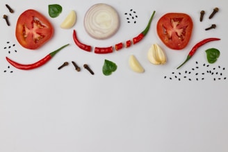 Slices of tomato and onion along with whole red chili peppers, garlic cloves, green leaves, black sesame seeds, and cloves are creatively arranged on a white background.