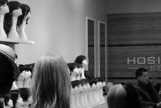 A shop interior with numerous mannequin heads displaying various wigs is visible. The neatly arranged display shows different hairstyles. A man appears to be seated toward the right, possibly resting or waiting.