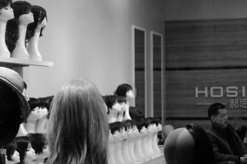 A shop interior with numerous mannequin heads displaying various wigs is visible. The neatly arranged display shows different hairstyles. A man appears to be seated toward the right, possibly resting or waiting.