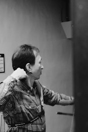 A person is pulling back a bow while aiming, wearing a plaid shirt. The environment appears to be indoors with minimalistic decor.
