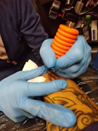 A person wearing blue gloves is holding a tattoo machine with an orange grip, applying ink onto a tattooed arm. The tattoo features intricate designs and the setting appears to be a tattoo studio.