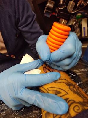 A person wearing blue gloves is holding a tattoo machine with an orange grip, applying ink onto a tattooed arm. The tattoo features intricate designs and the setting appears to be a tattoo studio.