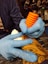 A person wearing blue gloves is holding a tattoo machine with an orange grip, applying ink onto a tattooed arm. The tattoo features intricate designs and the setting appears to be a tattoo studio.