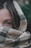 A close-up of a woman's face partially covered by a textured, plaid-patterned scarf, focusing on her eyes. The scarf is made of a wool-like material with a beige, black, and red checkered design. The background is slightly blurred, suggesting an outdoor setting with green foliage.