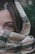 A close-up of a woman's face partially covered by a textured, plaid-patterned scarf, focusing on her eyes. The scarf is made of a wool-like material with a beige, black, and red checkered design. The background is slightly blurred, suggesting an outdoor setting with green foliage.