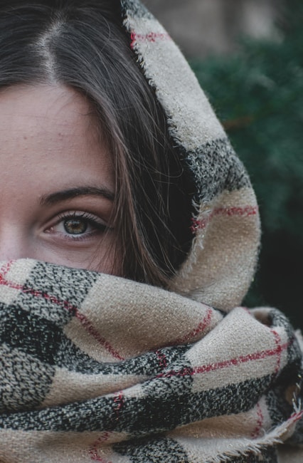 A close-up of a woman's face partially covered by a textured, plaid-patterned scarf, focusing on her eyes. The scarf is made of a wool-like material with a beige, black, and red checkered design. The background is slightly blurred, suggesting an outdoor setting with green foliage.