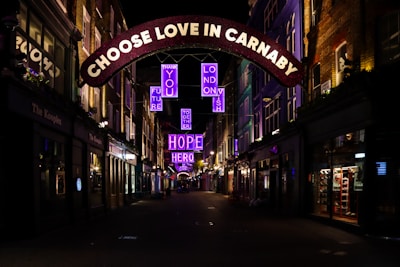 An illuminated street at night featuring vibrant neon signs suspended above, with messages of positivity such as 'CHOOSE LOVE IN CARNABY' and 'HOPE'. The street is bordered by buildings with shops, some of which have glowing lights inside.
