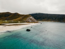 A serene coastal landscape with turquoise water gently lapping against a white sandy beach. A small cluster of quaint houses with red and white exteriors is nestled in the grassy hills. The hills rise dramatically with rugged textures, under a cloudy gray sky that gives a moody atmosphere.